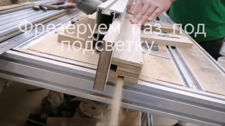 Мастерская: рука прижимает светлую деревянную планку к стопке таких же планок на станине станка, рядом лежит алюминиевый профиль, из-под заготовки вниз сыплется древесная пыль; поверх кадра надпись «Фрезеруем паз под подсветку»